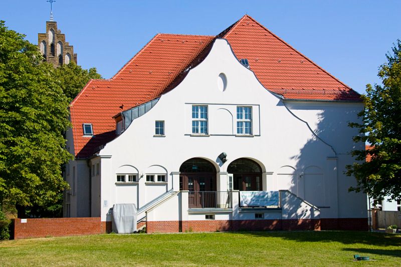Ein weißes Gebäude zwischen Bäumen und einem Kirchturm im Hintergrund