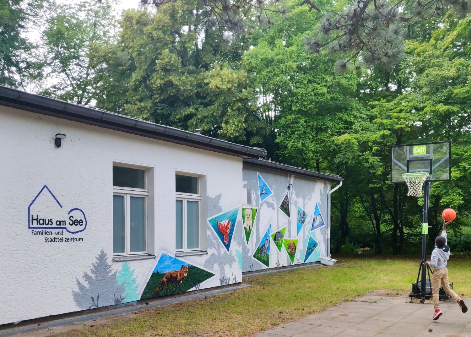 links Nebengebäude Haus am See mit Logo und Grafitti, rechts ein Junge, der einen Basketball in den Korb wirft