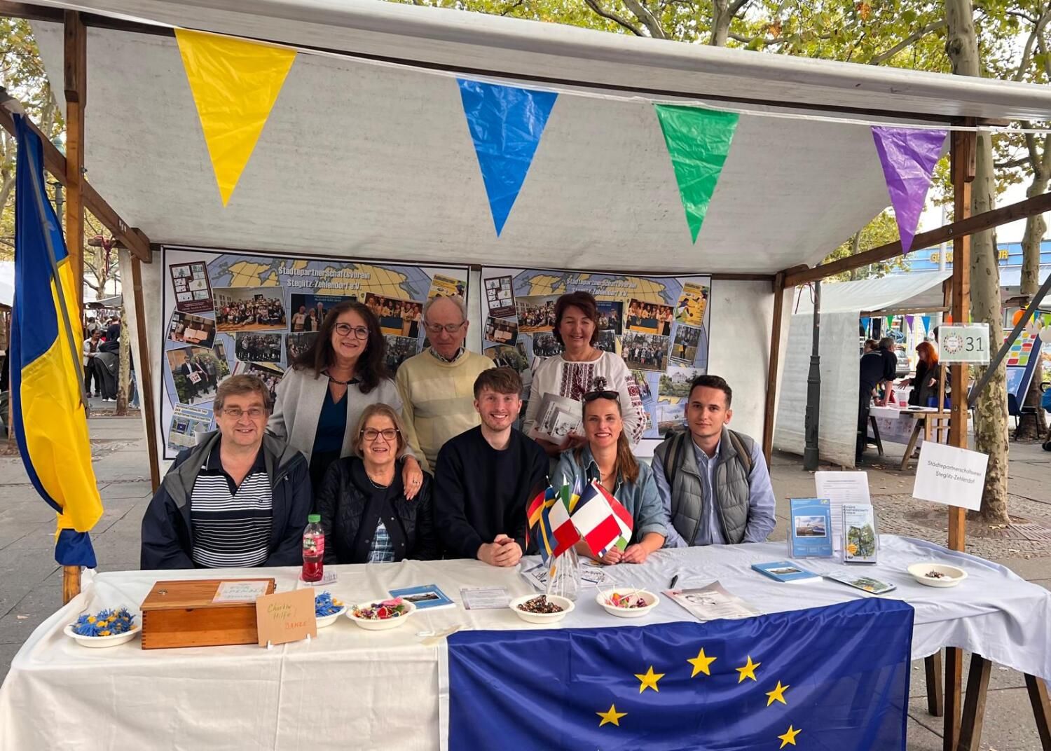 11. September 2026: Strahlen beim "Interkulturellen Fest" auf dem Hermann-Ehlers-Platz um die Wette: Mitglieder des Städtepartnerschaftsvereins Steglitz-Zehlendorf e.V. Erste Reihe Mitte: Vorsitzender Sebastian Leskien; zweite Reihe 1.v.l.: Vorstandsmitglied Olga Pischel; erste Reihe 2.v.l.: Gisela Pflug, langjährige Vereinsvorsitzende