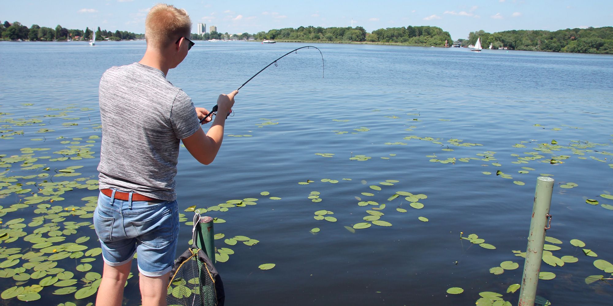 Angler. Ein sommerlich gekleiderter Angler steht auf einem Steg und hält eine Angelrute in einen See. Um den Steg herum sind Seerosenblätter. Im Hintergrund sieht man vor dem baumbestandenen gegenüberliegenden Seeufer einige Segelboote.