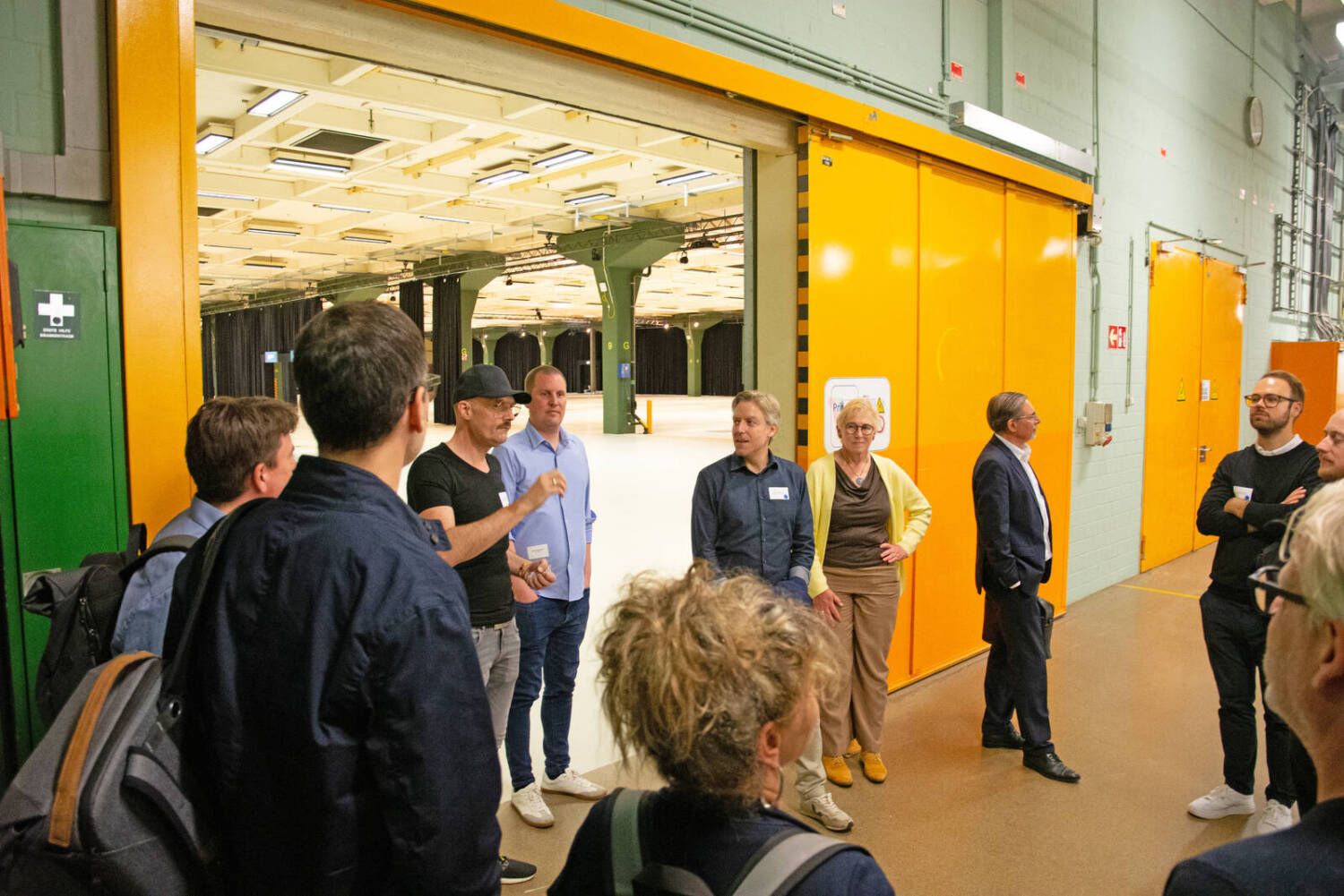 Gruppe von Personen in einem großen Raum mit geöffneter gelber Tür und leerem Hintergrund, einige tragen Namensschilder und hören einem Sprecher zu.