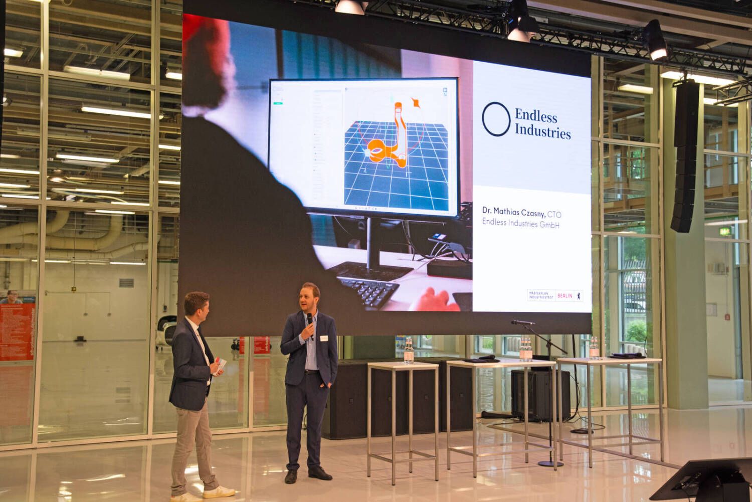 Zwei Männer stehen vor einer großen Leinwand mit einer Präsentation von Endless Industries, einer hält ein Mikrofon und spricht, während der andere zuhört.