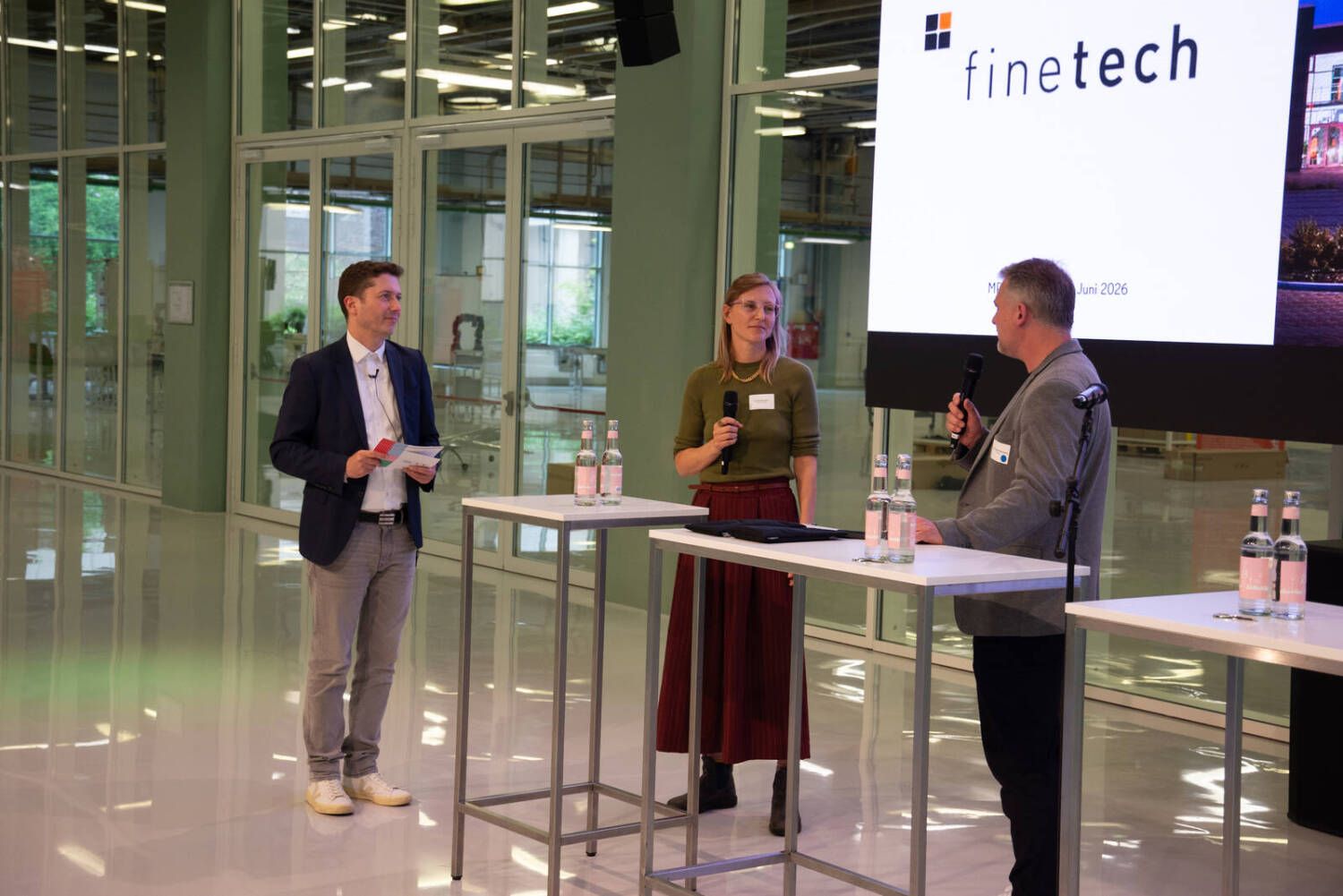Drei Personen stehen an drei nebeneinander aufgestellten Tischen vor einer großen Leinwand mit der Aufschrift finetech und sprechen in Mikrofone.