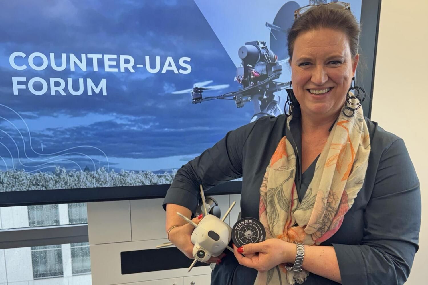 Frau hält zwei Drohnen vor einem Bildschirm mit der Aufschrift Counter-UAS Forum in der Hand