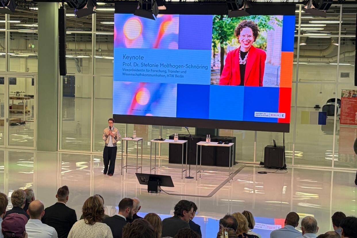 Eine Frau hält eine Keynote auf einer Bühne vor Publikum, hinter ihr ist eine Leinwand mit ihrem Porträt und Informationen zur Veranstaltung.