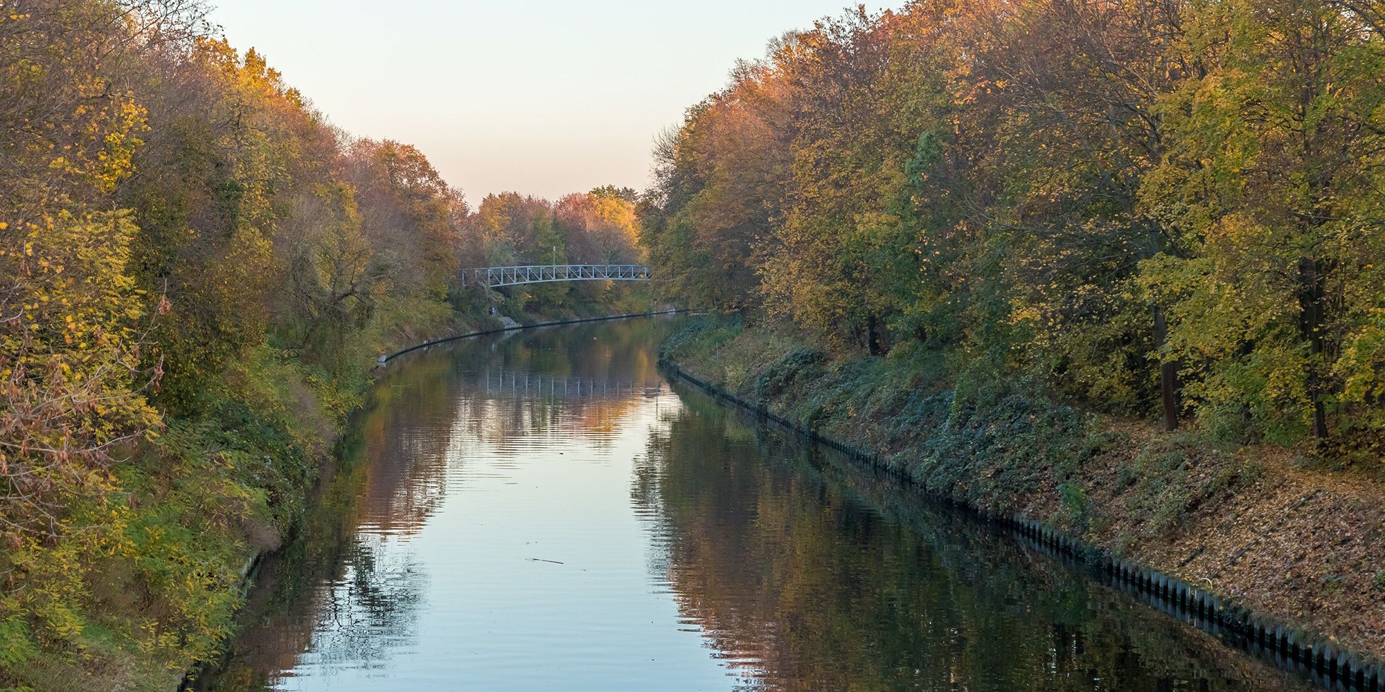 Das Foto zeigt einen etwas erhöhten Blick auf den Teltowkanal. Herbstlich gefärbte Bäume säumen ihn zu beiden Seiten. Etwas entfernt überspannt eine Brücke das ruhige Gewässer. An dieser Stelle macht der Wasserlauf eine langgezogene Biegung nach rechts. Verschwommen reflektiert die leicht gekräuselte Wasseroberfläche die dichte Ufervegetation in Gelb-, Rot- und Grüntönen sowie den blassblauen Himmel.