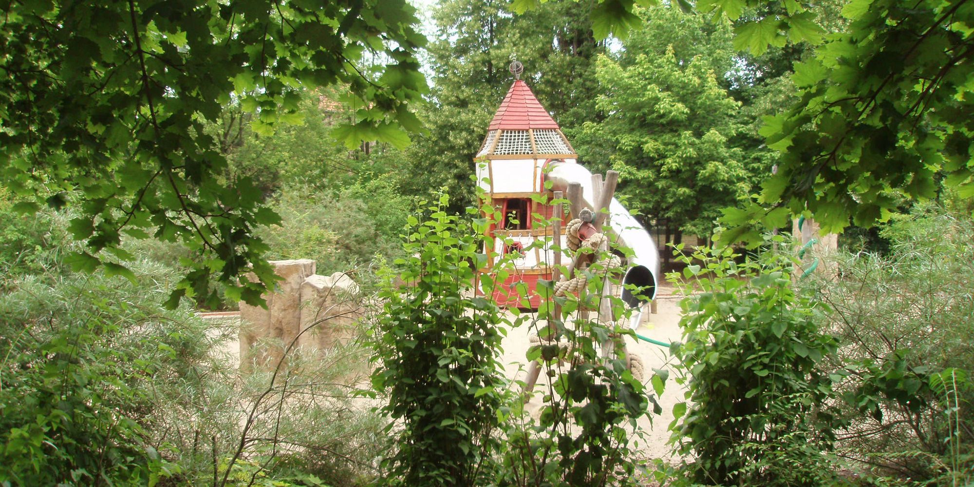 Der Spielplatz der Reinhardswald-Grundschule im Bezirk Friedrichshain-Kreuzberg ist von grünen Büschen, Bäumen und Sträuchern umgeben. Inmitten einer Sandfläche ragt ein Kletterturm mit rotgestrichener Spitze in die Höhe. Eine Röhrenrutsche aus Edelstahl windet sich von dem eckigen Spielhaus zu Boden. Links stehen zwei Natursteinblöcke im hellgelben Pudersand.