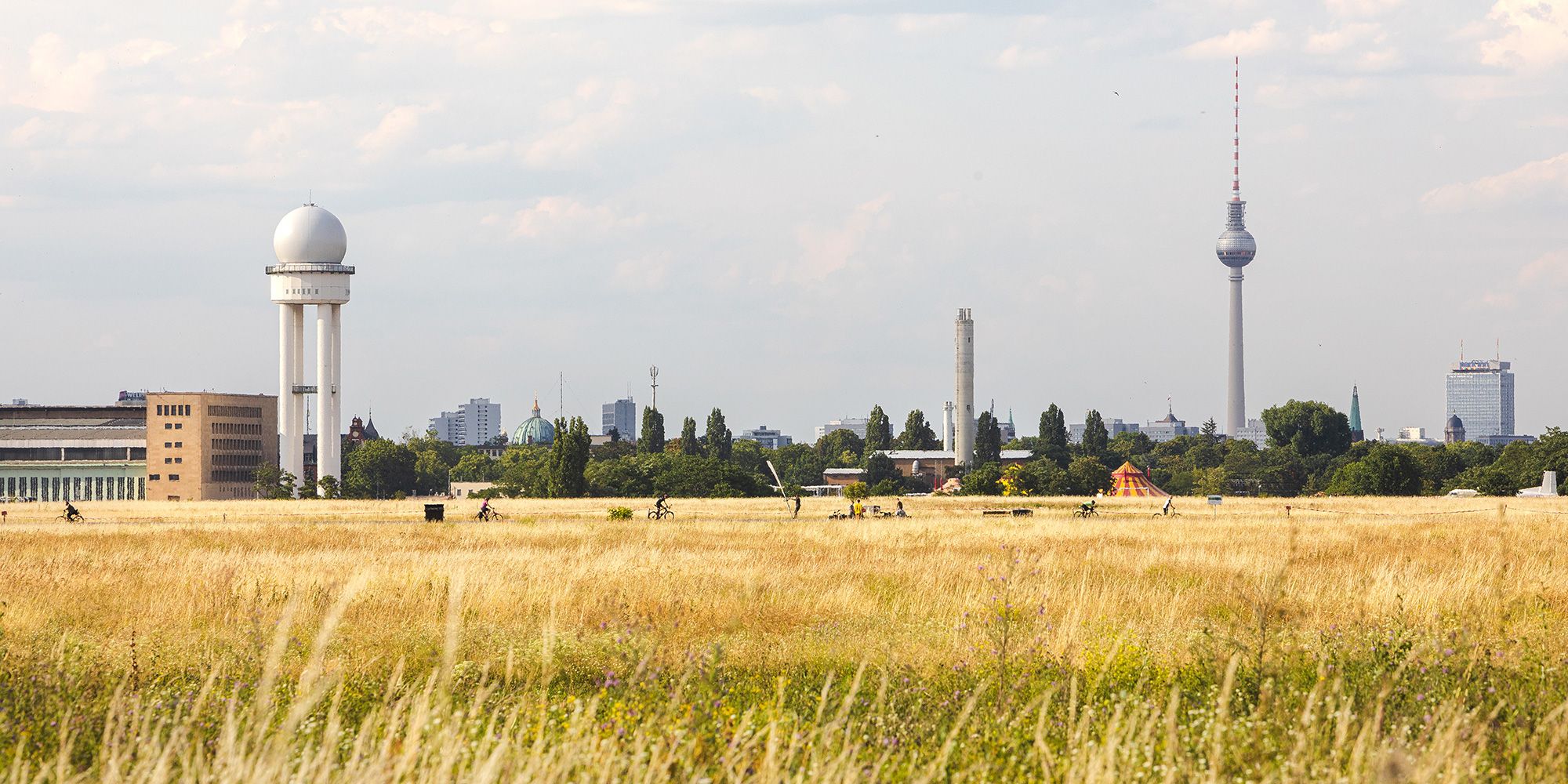 Das Panoramafoto zeigt einen Blick über das sogenannte Wiesenmeer aus gelben Ähren und Gräsern auf dem Tempelhofer Feld. Die weitläufige Freifläche nimmt etwa das untere Drittel des Bildes ein. In großer Entfernung sind einige Radfahrer auszumachen. Hinter grünen Baumkronen im Hintergrund die Skyline von Berlin. Von links nach rechts: ein Gebäude des ehemaligen Flughafens und der massive Radarturm mit kugelförmiger Kuppel, einige Hochhäuser, die Kuppel des Berliner Doms sowie der Fernsehturm.