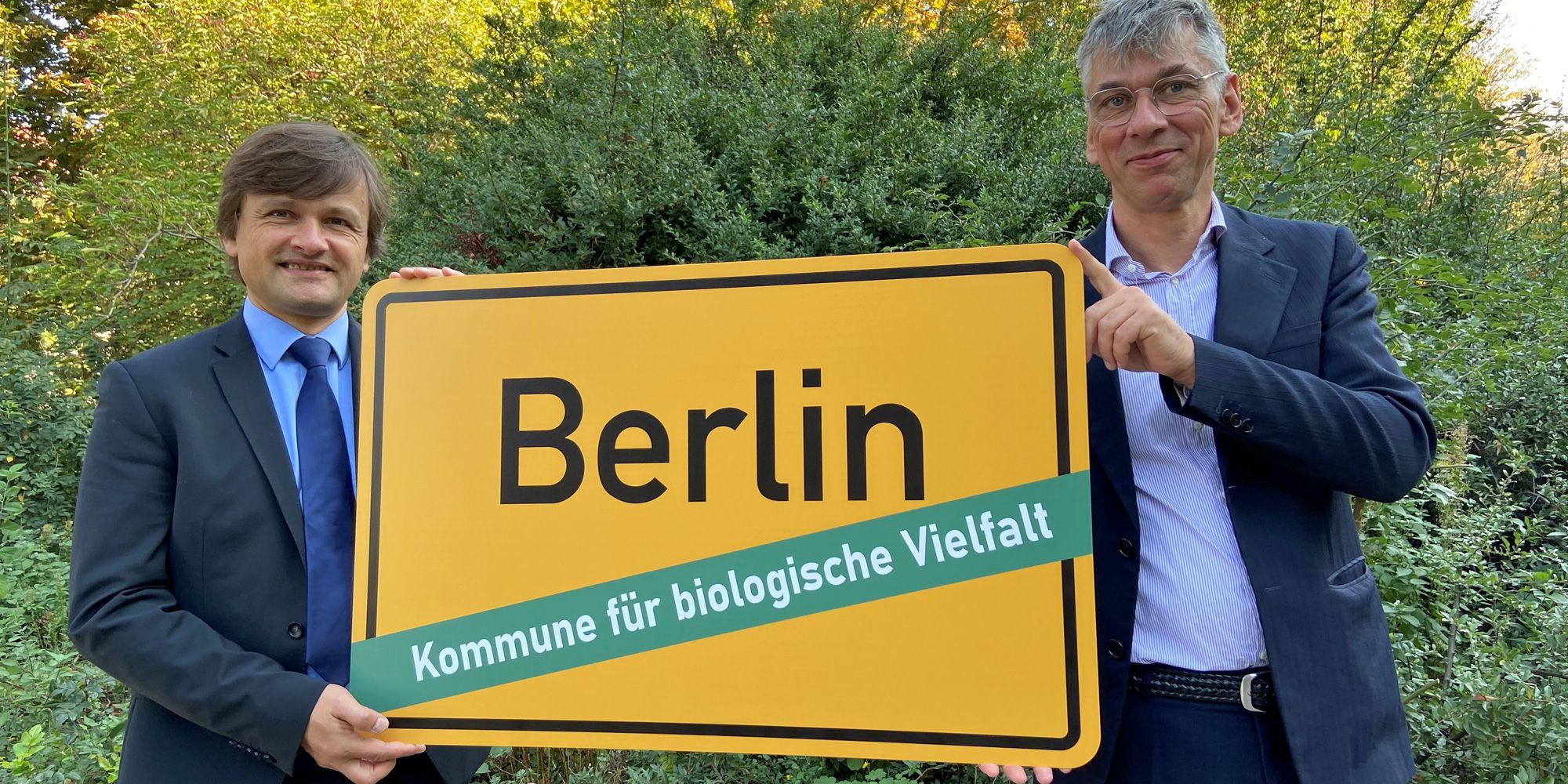 Vor grünen Büschen halten Robert Spreter und Staatssekretär Stefan Tidow in Hemd und Jackett das gelbe Berliner Ortseingangsschild hoch. Unter dem Stadtnamen steht: Kommune für biologische Vielfalt. Anlass ist der Beitritt Berlins dem Bündnis am 1. Oktober 2020.