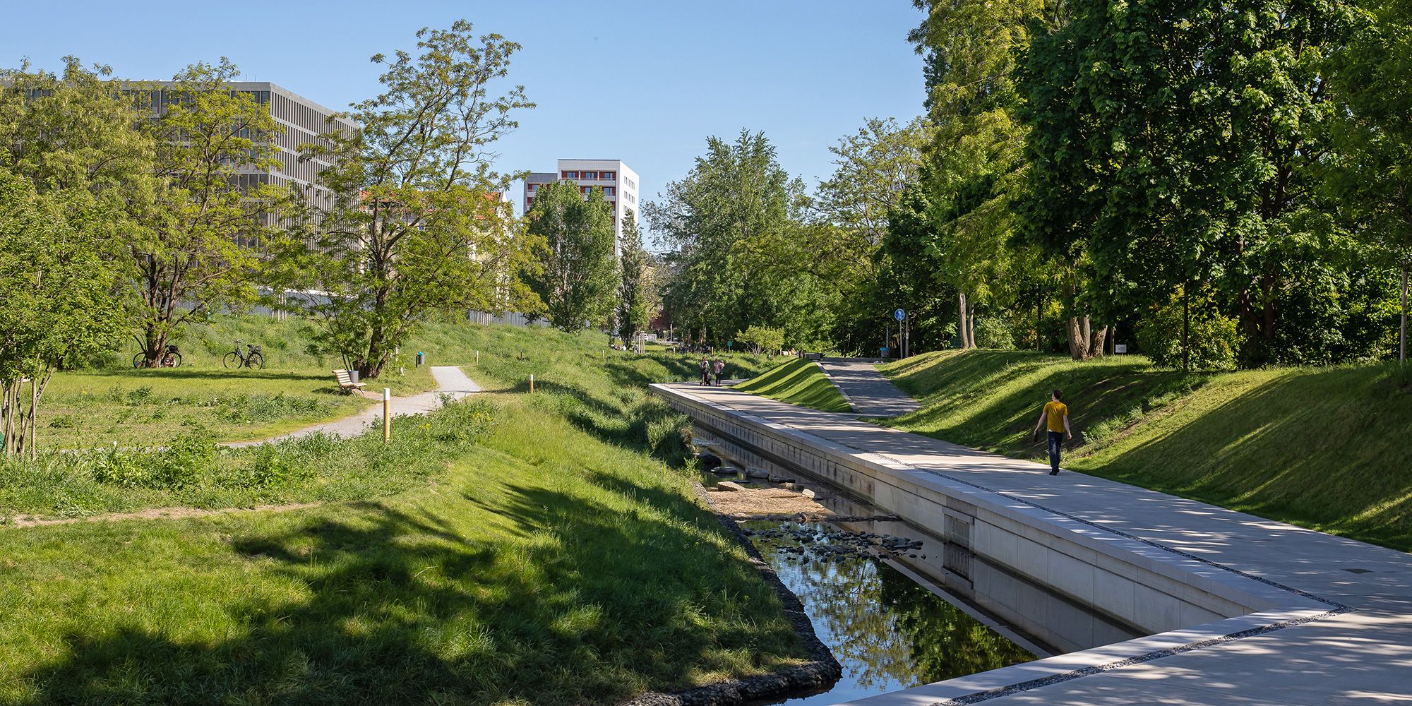 Eine parkähnliche Grünanlage mit Bäumen und Wiese durchzieht das Bild. Ein schmaler Bach fließt in der Mitte, daneben verläuft ein breiter, gepflasterter Weg, auf dem eine Person spaziert. Links sind moderne Gebäude und Sitzbänke zu sehen, rechts dichter Baumbestand. Der Himmel ist klar und blau.