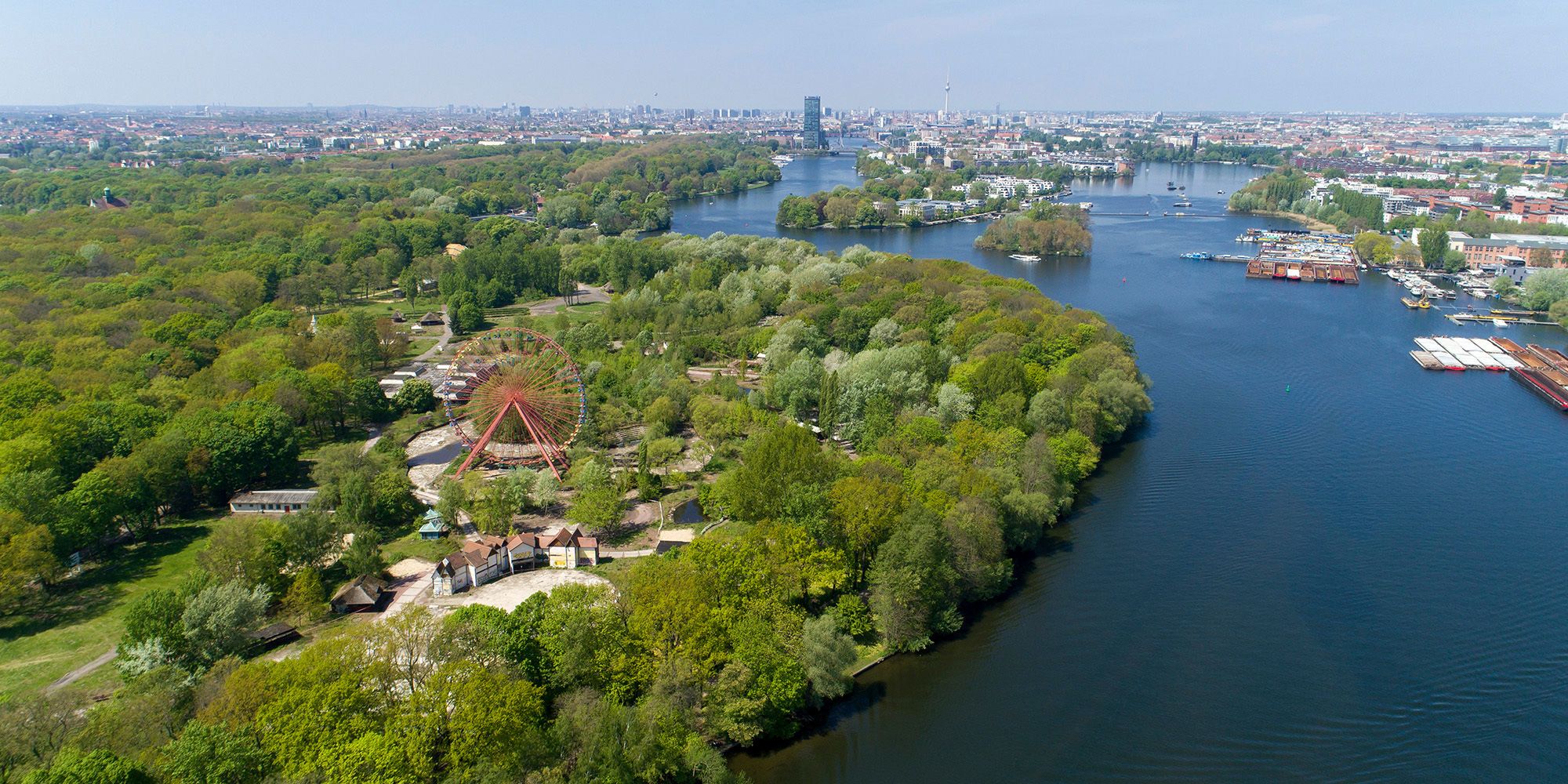 Aus großer Höhe ein Blick auf den ausgedehnten grünen Plänterwald, der sich links im Bild parallel zur tiefblauen Spree erstreckt und in den Treptower Park übergeht. Im Vordergrund ragt zwischen Bäumen das rote Riesenrad des ehemaligen Spreeparks auf. In weiter Ferne lassen sich auf der Spree die Halbinsel Stralau und die Rummelsburger Bucht erkennen. Das markante Hochhaus der Treptowers zeichnet sich vor dem blauen Himmel ab.