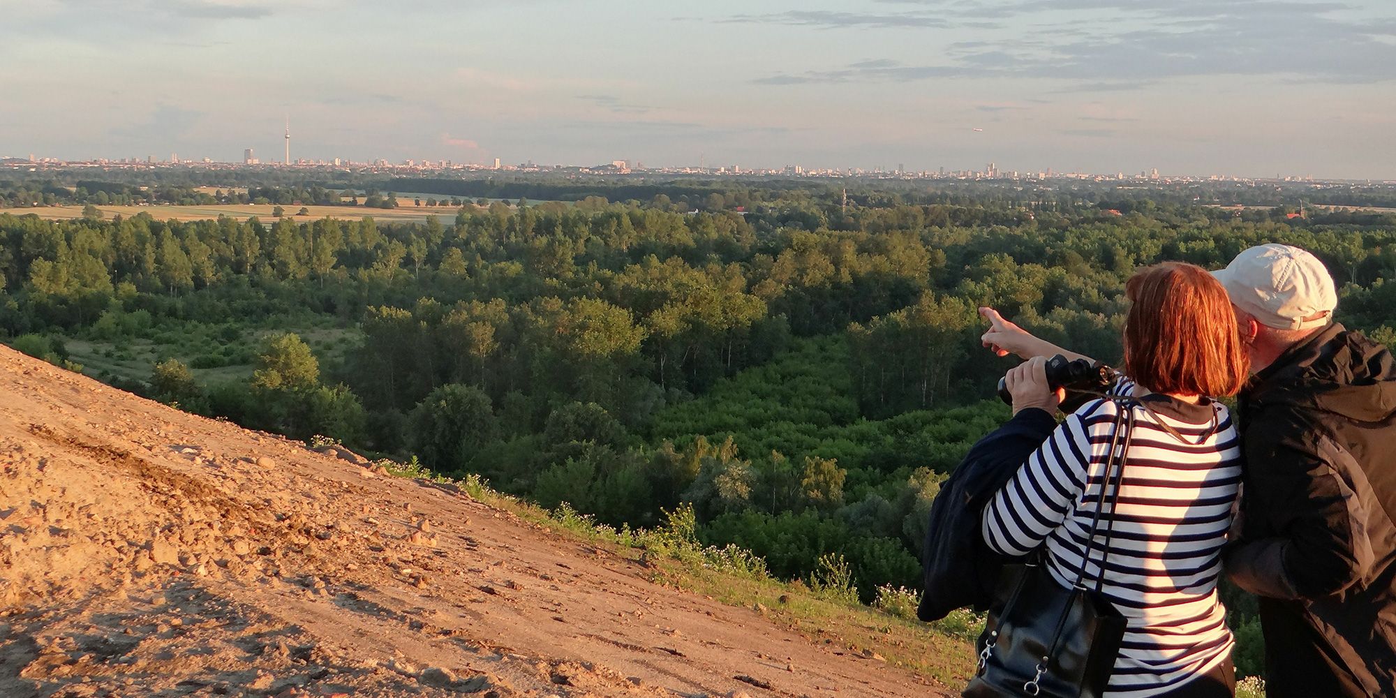 In der Abendsonne stehen am rechten unteren Bildrand abgewandt eine ältere Frau und ein Mann auf einem Sandweg. Sie schauen von einem Hügel auf eine Landschaft mit Laubwäldern und Wiesen. Am Horizont zeichnet sich die Silhouette von Berlin ab. Der blaue Himmel wird von einigen Wolkenbändern durchzogen. Die Frau hat halblanges, rotes Haar und trägt ein schwarz-weiß geringeltes Shirt. Sie deutet in die Ferne und hält ein Fernglas. Der Mann trägt ein beiges Käppi und eine schwarze Jacke.