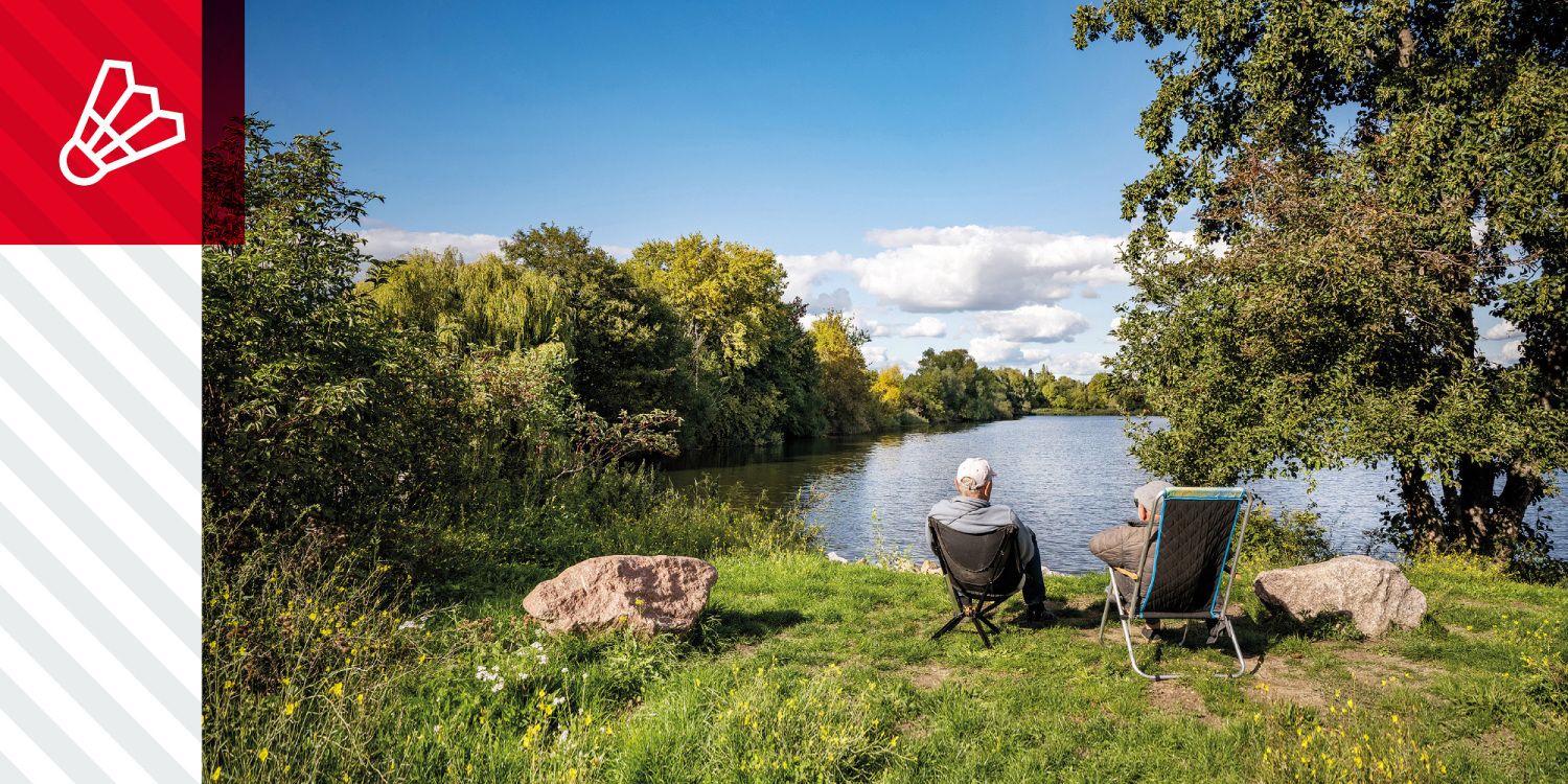 Zwei Menschen sitzen auf Stühlen am Ufer eines Flusses, umgeben von Bäumen und Wiese.