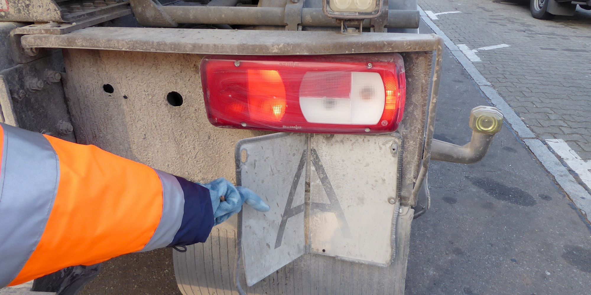 Ein Arm in Warnjacke hält ein Schild an das Heck eines Fahrzeugs, auf dem ein „A“ zu sehen ist. Dies zeigt an, dass in dem Fahrzeug Abfälle nach deutschem Recht transportiert werden.