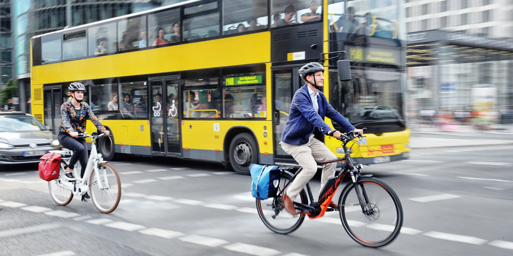 Zwei E-Bikes fahren auf einem Radweg. Dahinter fährt ein gelber Bus. Im linken Rand des Bildes sieht man die Kühlerhaube eines fahrenden Autos.