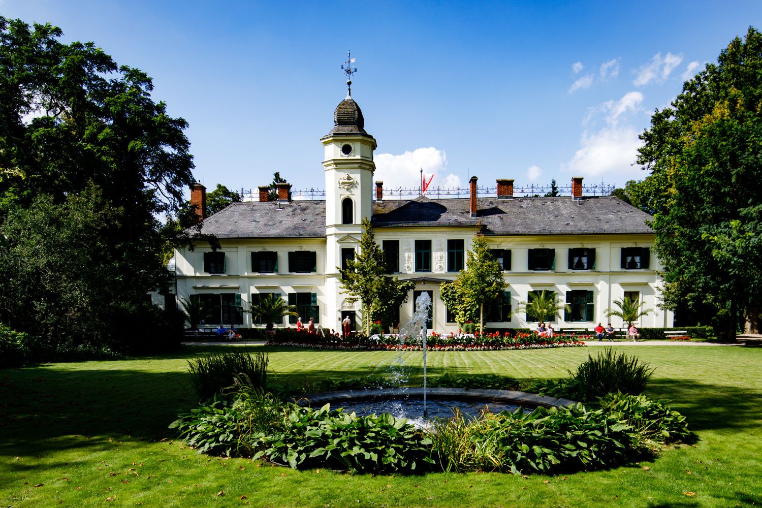 Im Vordergrund eine grüne Wiese. Im Hintergrund das Schloss Britz. Ein weißes Gebäude mit grünen Fensterläden. In der Mitte des Hauses ein schmales Türmchen. Rechts und links des Bildausschnittes belaubte Bäume