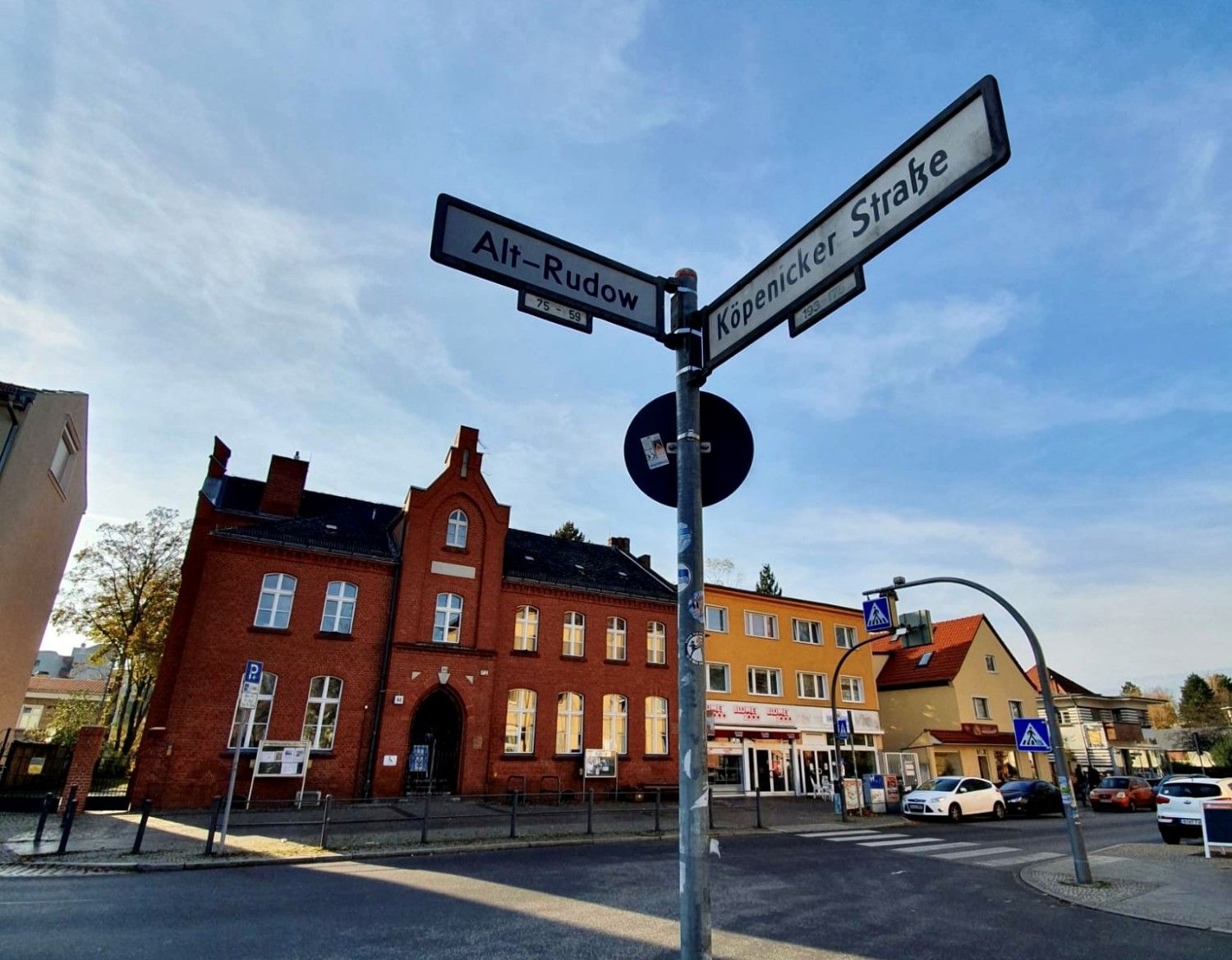 Im Vordergrund eine Straßenkreuzung. Auf der Ecke steht ein Straßenschild. Nach links zeigt das Schild "Alt-Rudow" nach rechts das Schild "Köpenicker Straße". Im Hintergrund ein rotes Backsteinhaus- die alte Dorfschule Rudow. Daneben ein Haus mit Flachdach in orange. Der Himmel ist blau mit leichten Schleierwolken.