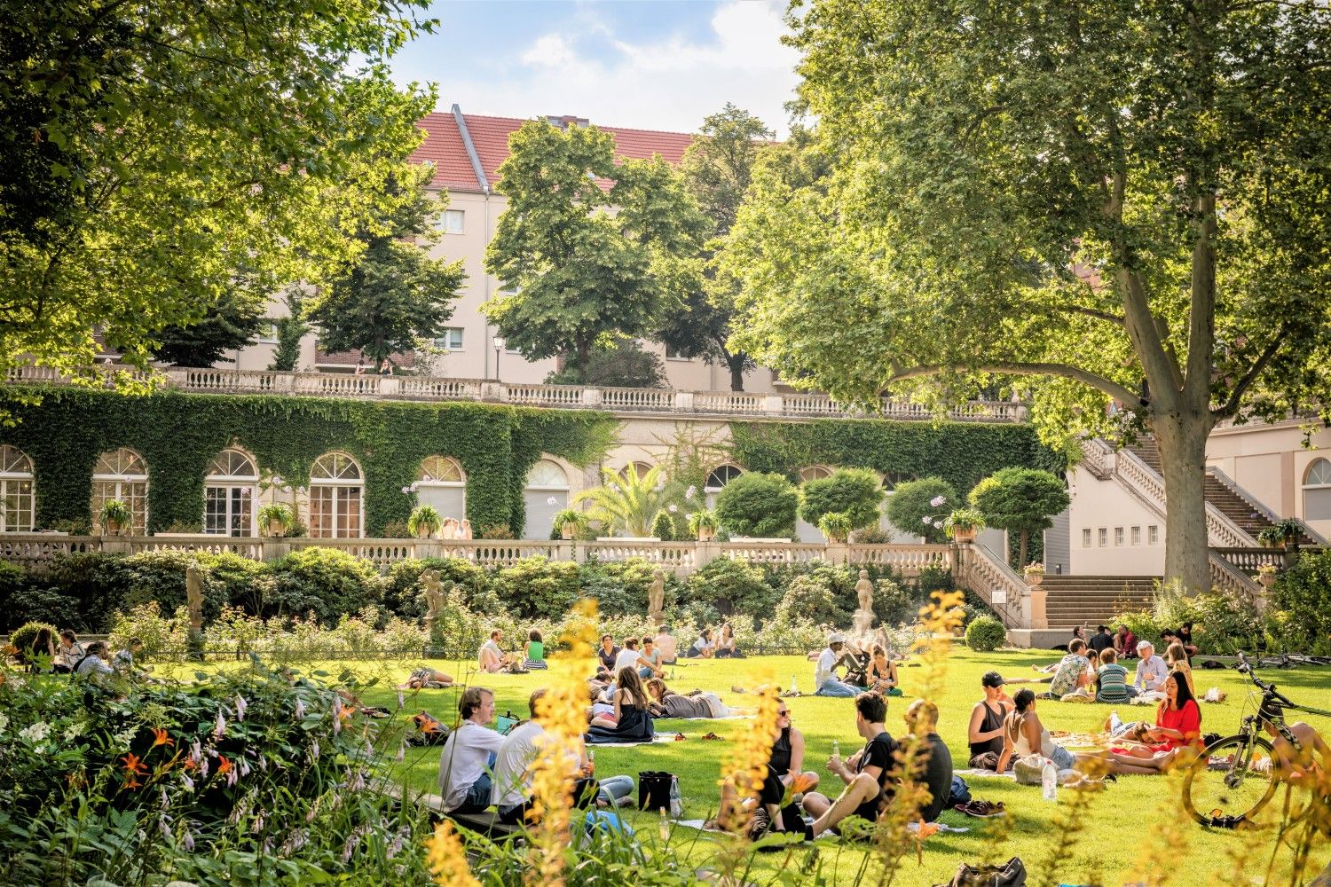 Menschen sitzen in kleinen Gruppen auf einer Wiese. Rechts sieht man einen Baum. Im Hintergrund ein Laubengang. Die Sonne scheint