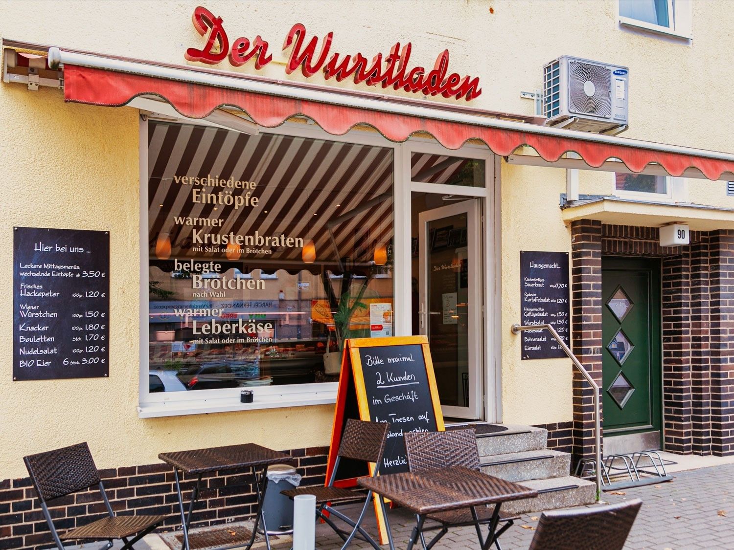 Gelbe Hausfassade mit einem großen Schaufenster und daneben einer geöffneten glässernen Eingangstür. Drei Stufen führen zu dem Ladengeschäft hoch. Links von den treppen steht eine Aufstelltafel. Davor zwei Tische mit jeweils zwei Stühlen. Über dem Schaufenster ein roter verschlungener Schriftzug "Der Wurtsladen"
