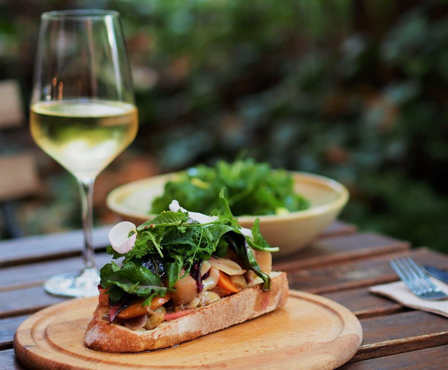 Auf einem dunklen Tisch steht ein Weinglas mit Weißwein und auf einem hölzernen rett liegt eine Scheibe Baguette mit Salat drauf