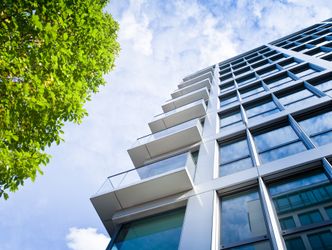Blick von unten auf ein modernes Hochhaus mit Glasfassade und Balkonen neben einem Baum mit grünen Blättern