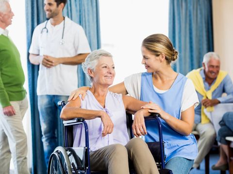 Eine Pflegerin hockt neben einer Frau im Rollstuhl und hat ihre rechte Hand auf die Schulter der Person im Rollstuhl gelegt. Sie lächelt sie an.
