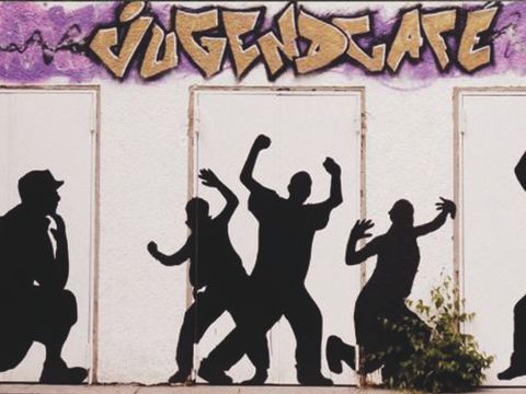 Schwarze Figuren, die tanzen mit Graffiti an der Wand im Hinergrund mit dem Text: Jugendcafe