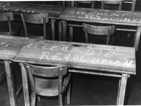 Bildvergrößerung: Hier sehen Sie ein schwarz weiß Foto mit Holztischen und Stühlen, auf dem einen Tisch steht mit Kreide geschrieben: Es lebe die Revolution