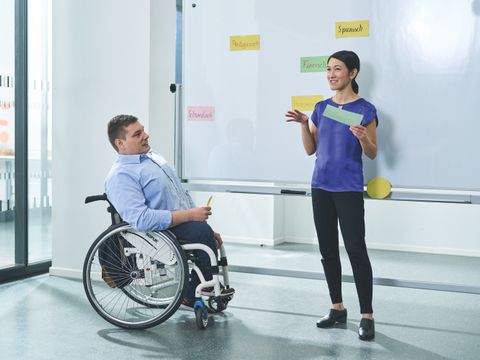 ein Mann im Rollstuhl mit einer Lehrerin vor einem Whiteboard im Unterricht