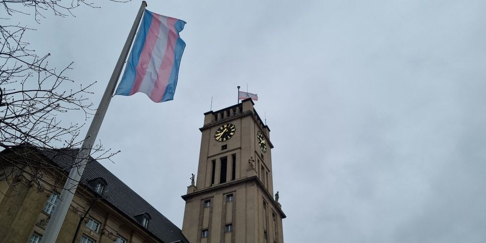 Vor einem Gebäude mit Uhrenturm weht eine blau-rosa-weiß gestreifte Fahne an einem Fahnemast.
