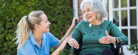 Eine Pflegerin hockt neben einer Frau im Rollstuhl auf einer Terrasse. Beide lächeln sich zufrieden an.