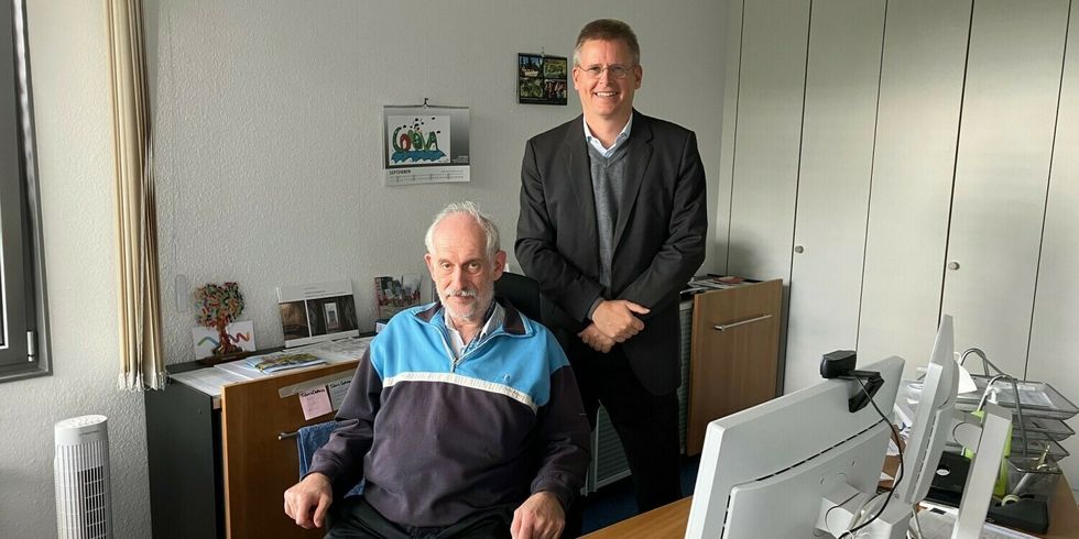 Zwei Männer sind in einem Büro. Ein Mann sitzt auf einem Drehstuhl vor einem Arbeitsplatz mit Computer. Der andere Mann steht hinter ihm.