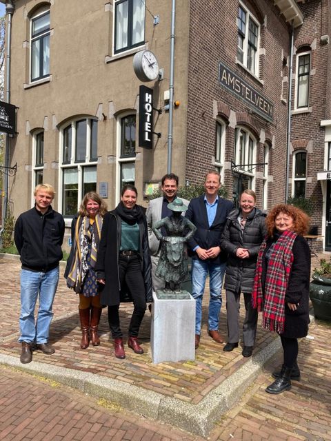 Bildvergrößerung: Sieben Personen stehen nebeneinander hinter einer Statue vor einem Hotel mit dem Textzug "Amstelveen".