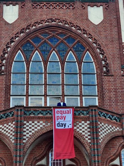 Bildvergrößerung: Bezirksbürgermeister Oliver steht auf dem Balkon des Rathauses, an dem die Fahne zum Equal Pay Day hängt.
