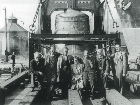 Enlarge photo: Six men and one woman in front of a large bell are smiling at the camera.