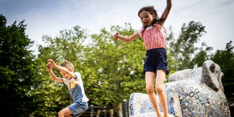 Ein Mädchen und ein Junge springen von einem mit Mosaiken verzierten Spielgerät. Das Mädchen trägt ein rot-rosa-weißes Top und blaue kurze Hosen. Der Junge trägt eine knielange Jeans und ein T-Shirt mit einem weißen, gelben und blauen Streifen. 