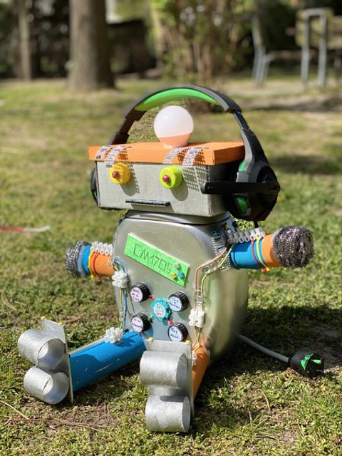 Bildvergrößerung: Ein selbst gebastelter Roboter in Alu-Optik sitzt auf der Wiese und hat Kopfhörer aus.