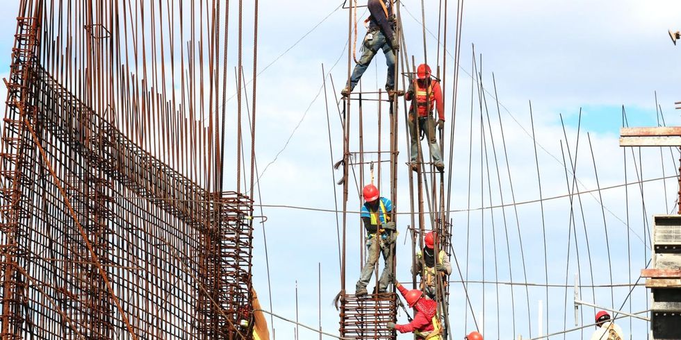 Arbeiter*innen bauen ein Gerüst auf in großer Höhe auf einer Baustelle im Freien.