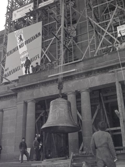 Enlarge photo: A large bell hangs on a rope in front of large building. A sign with the inscription “Berlin Emergency Program with Marshall Plan Aid” hangs on a scaffold in front of the building. 