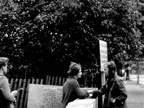 Bildvergrößerung: Zwei Mädchen an der noch nicht fertiggestellten Berliner Mauer in Treptow in der Harzer Straße. Sie sprechen miteinander und fassen sich an. Etwas weggedreht steht ein Soldat. Er hat keine Pistole und kein Gewehr in der Hand, sondern eine Blume.