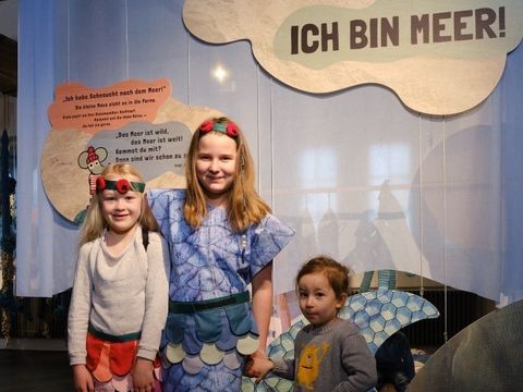 Bildvergrößerung: Drei Kinder in Kostümen stehen vor einem blauen Vorhang mit dem Schriftzug "Ich bin Meer!"
