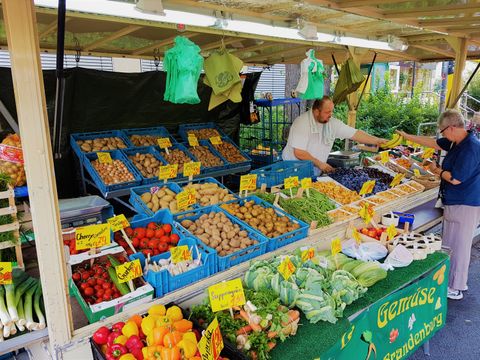 Obst und Gemüse Marktstand. Auf der linken Seite des Standes wird Gemüse verkauft: Paprika, Kohl und viel Kartoffeln. Auf der rechten Seite wird Obst verkauft: Äpfel und Pflaumen. Das Obst und Gemüse ist sortenrein in einzelnen Kästen aufgebaut. Hinter dem Marktstand steht ein Mann in einem weißen T-Shirt. Auf der anderen Seite des Marktstands steht ein Mann in einem blauen T-Shirt und einer hellen Hose, der dem Verkäufer gerade mehrere Bananen herüber reicht. 