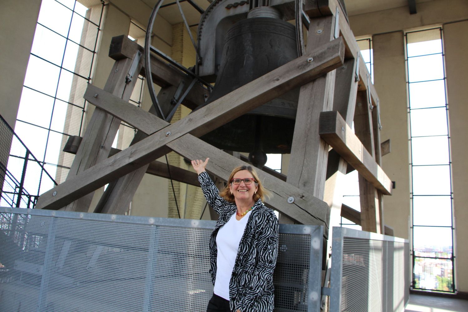 Smiling woman is standing in front of a large cast iron bell, which is hanging from a wooden scaffolding.