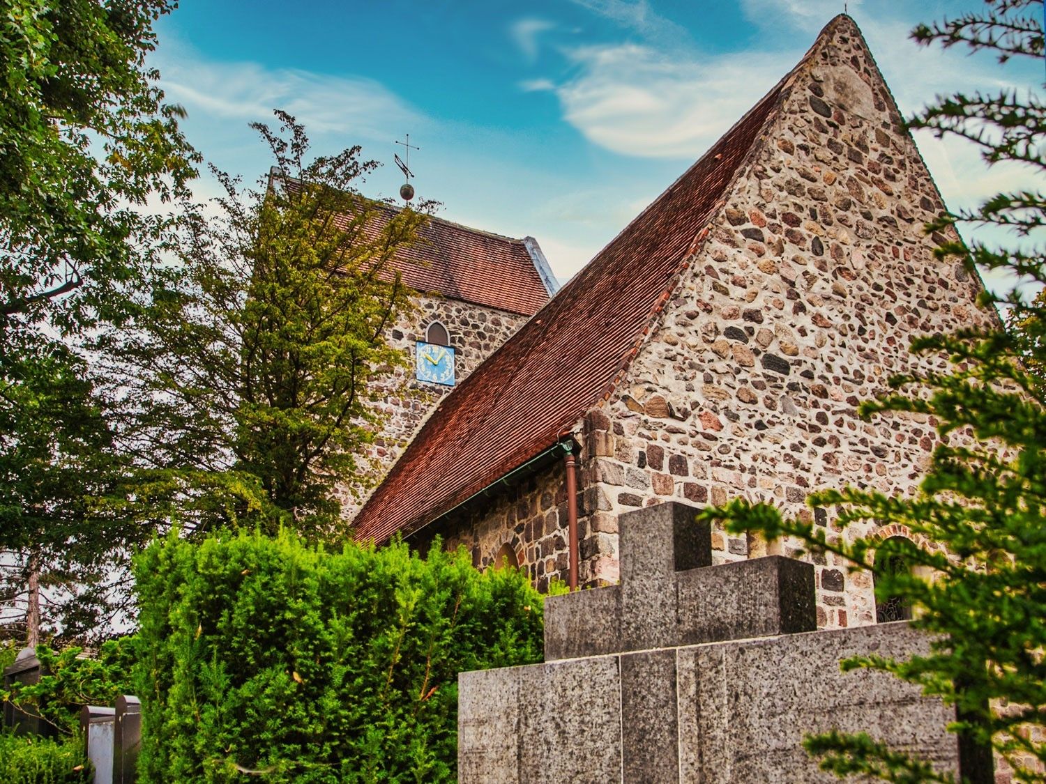 alte Dorfkirche in Buckow. Die Kirche besteht aus Feldsteinen in grau/braun mit einem roten Dach. Rechts und links von der Kirche Bäume und Sträucher.