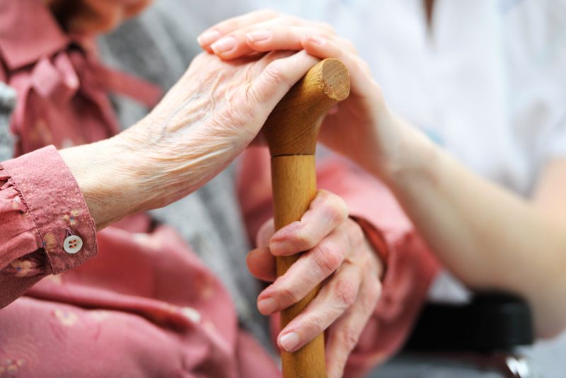 Eine alte Frau hält sitzend einen Gehstock mit der linken Hand umfasst und hat die rechte Hand auf den Griff gelegt. Darüber ist die Hand einer jüngeren Pflegeperson zu sehen.
