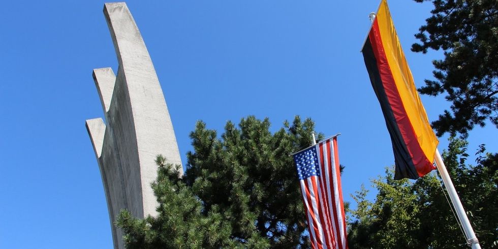 Unter einem klaren blauen Himmel sieht man das Luftbrückendenkmal, eine Gebilde aus Beton. Im Vordergrund sieht man die Amerikanische und die Deutsche Flagge an zwei Fahnenmästen hängen.