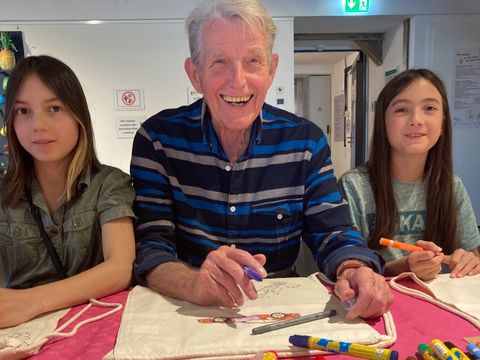 Bildvergrößerung: Ein älterer Mann sitzt zusammen mit zwei Mädchen an einem Tisch. Sie malen mit Stiften Beutel an.