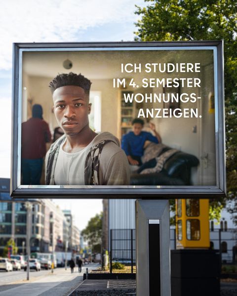 Auf einer Werbetafel in Berlin ist ein junger Mann abgebildet, der direkt in die Kamera blickt. Hinter ihm sind weitere Personen in einem Raum. Der Text auf der Tafel lautet: “ICH STUDIERE IM 4. SEMESTER WOHNUNGSANZEIGEN.” Im Hintergrund sieht man städtische Gebäude und Straßen.