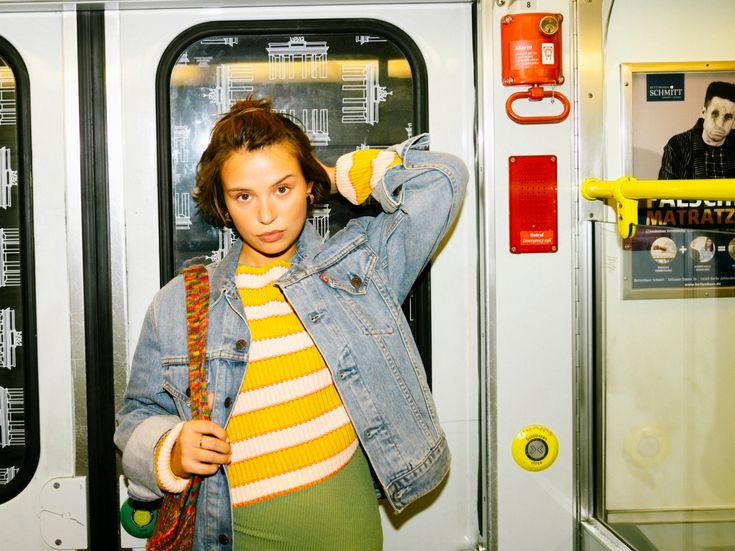 Eine junge Frau mit kurzen braunen Haaren steht in einem U-Bahn-Waggon. Sie trägt eine bunte gestreifte Strickjacke und eine Jeansjacke. Sie schaut direkt in die Kamera und hält eine Hand am Kopf. Im Hintergrund sind die Türen der U-Bahn und verschiedene Anzeigen zu sehen.