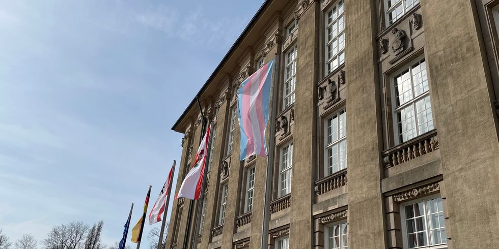 Eine blau-rosa-weiß gestreifte Fahne hängt an einem Fahnenmast neben mehreren anderen Mästen vor einem großen Gebäude.